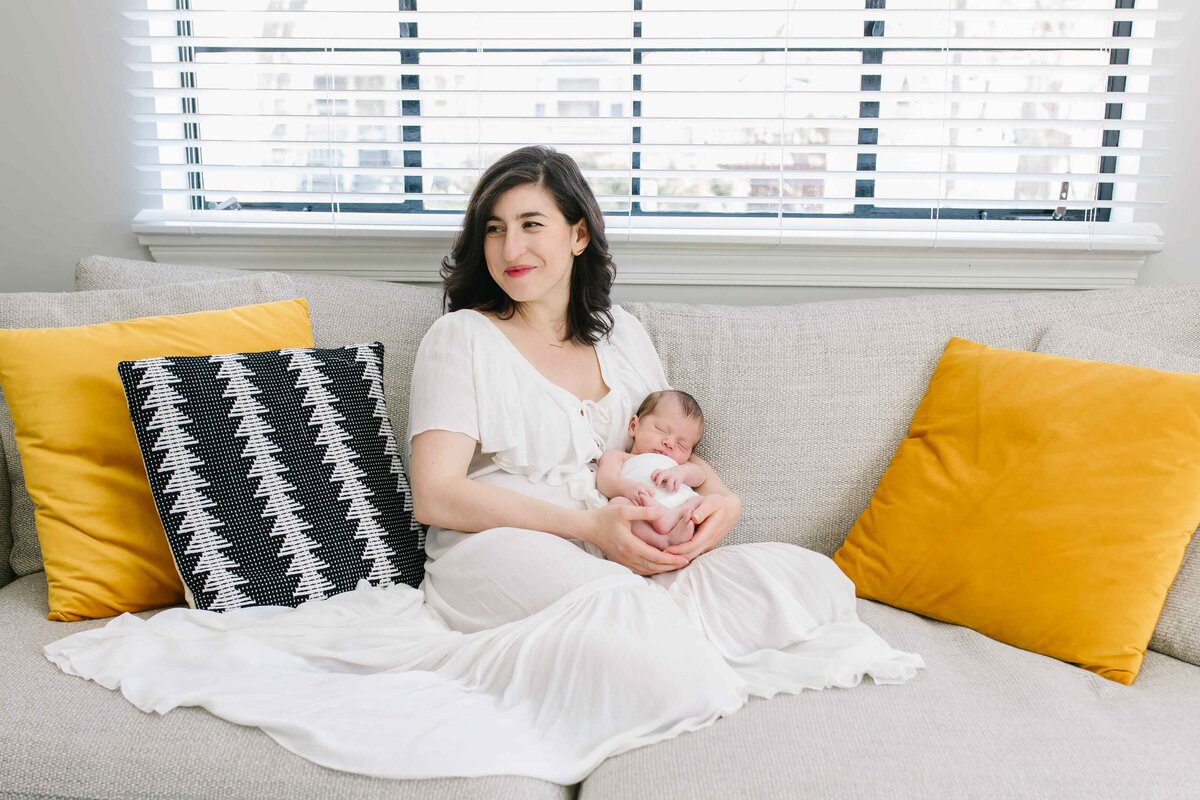 mother in white dress sitting on couch holding newborn baby