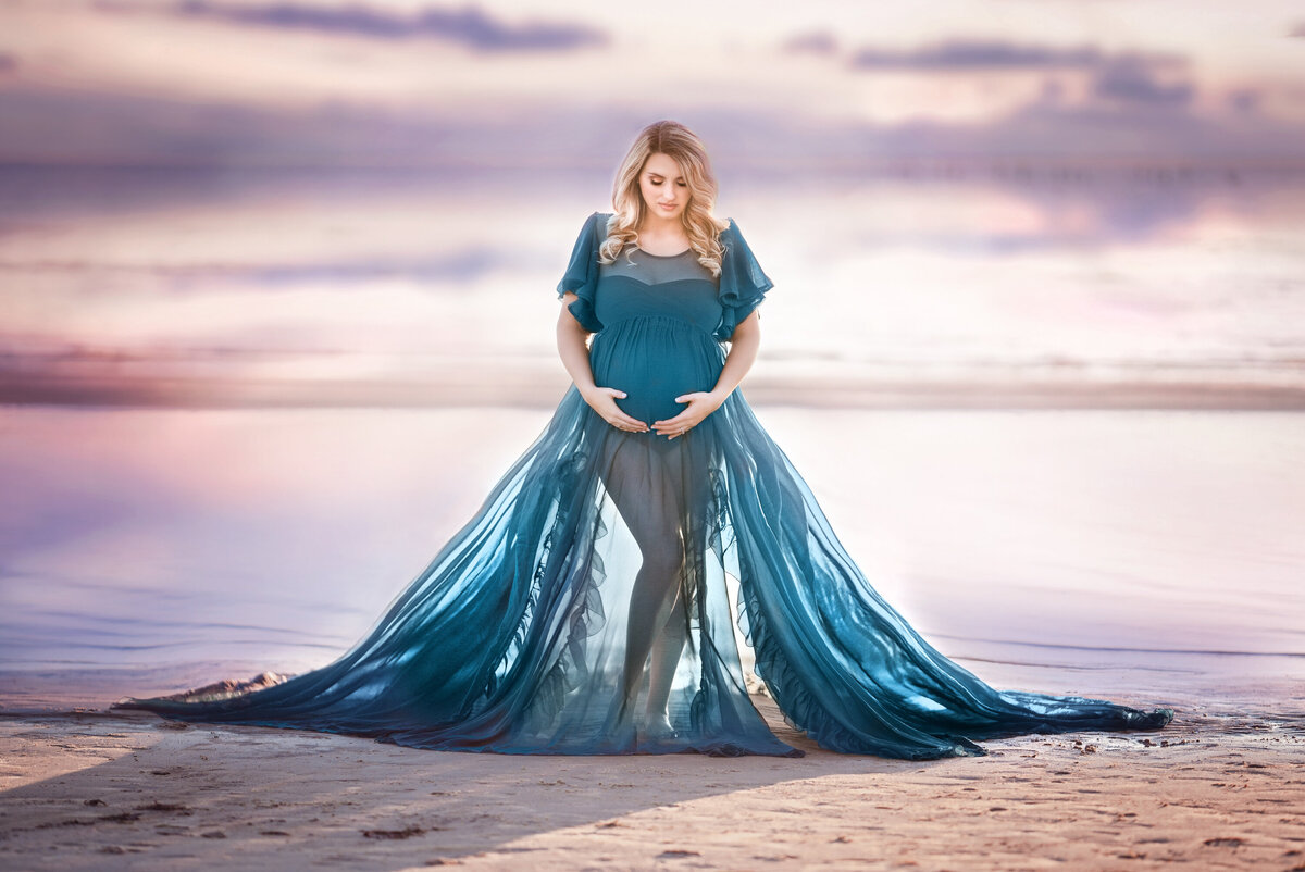 Blonde women with long, wavy hair standing on the beach at Fontainebleau State Park.  She is wearing a sheer marine blue dress and is very pregnant.  There is a lavender and pink sunset behind her.