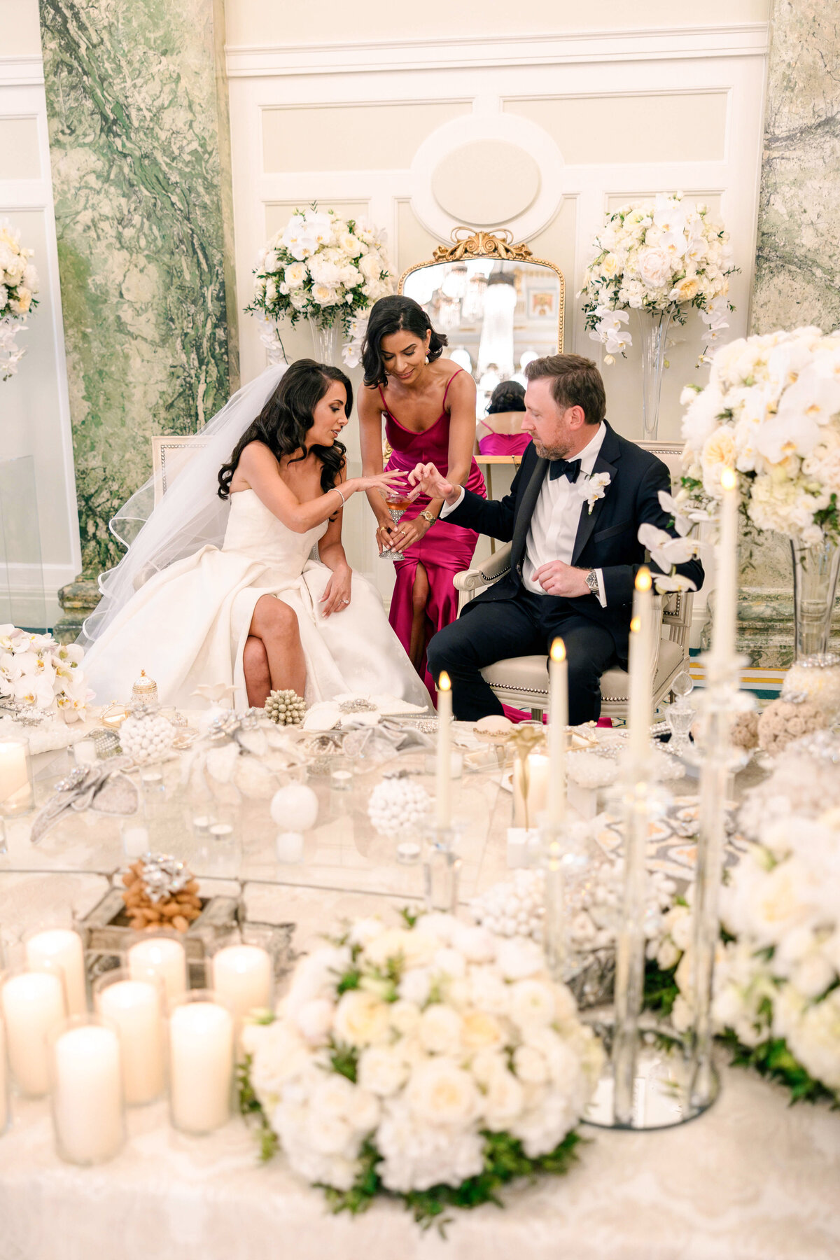 A bride and groom sit at a wedding ceremony table adorned with white flowers and candles. A woman in a red dress assists them with a ritual. The setting is elegant with marble walls and bouquets in the background.