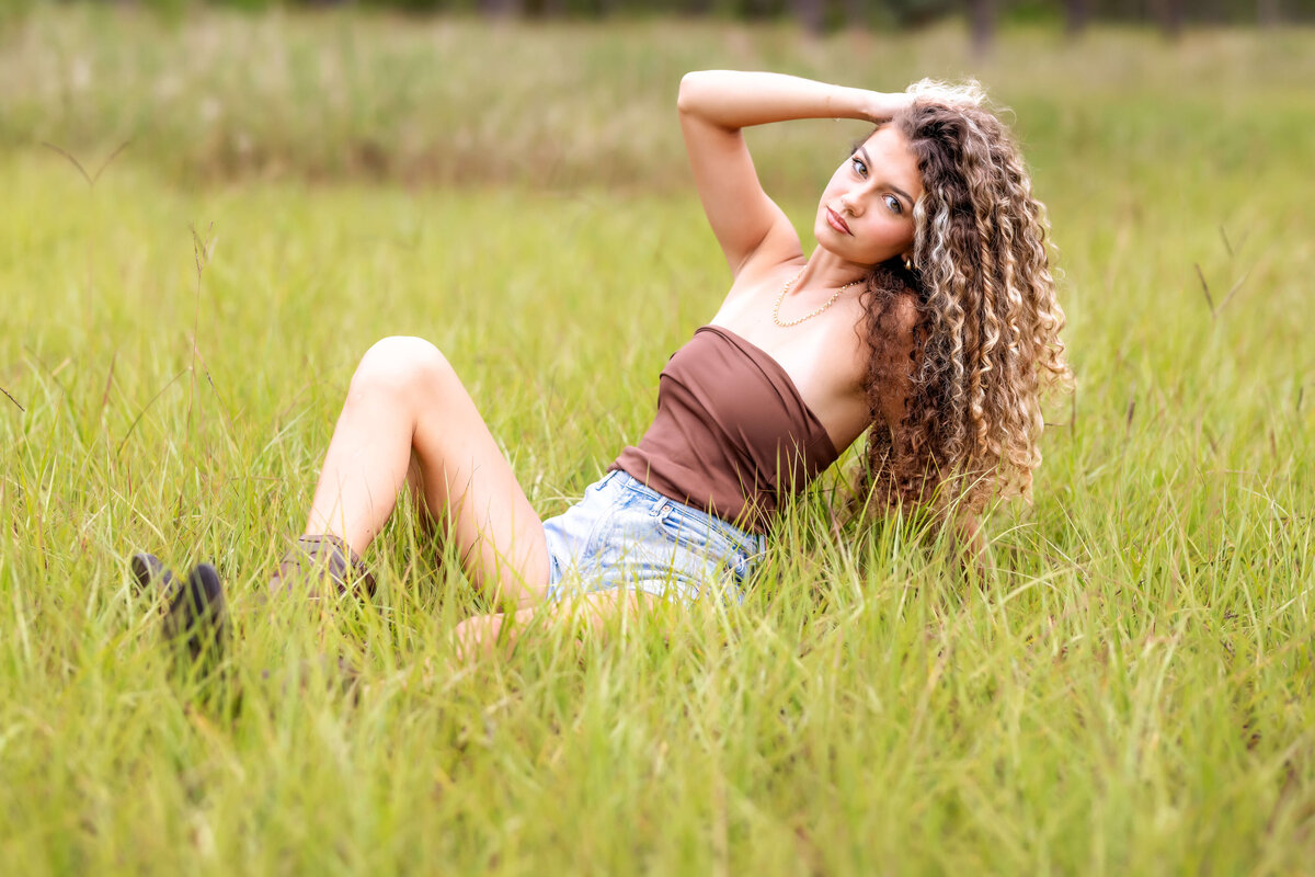 Orlando senior pictures featuring a teen girl sitting in tall green grass during sunset.