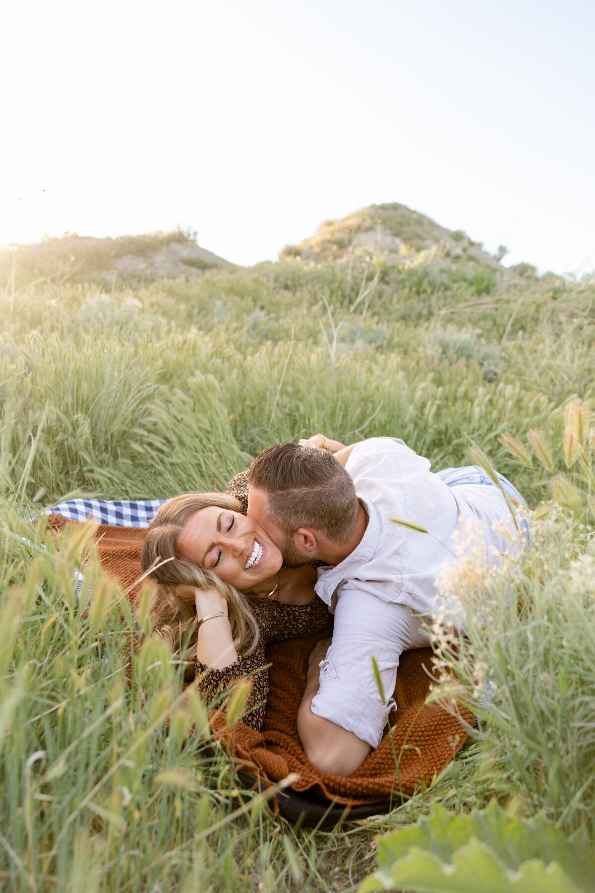 A couple laughs together as they lie in the grass, wrapped in a cozy blanket during their engagement session in Saratoga Springs, Utah.