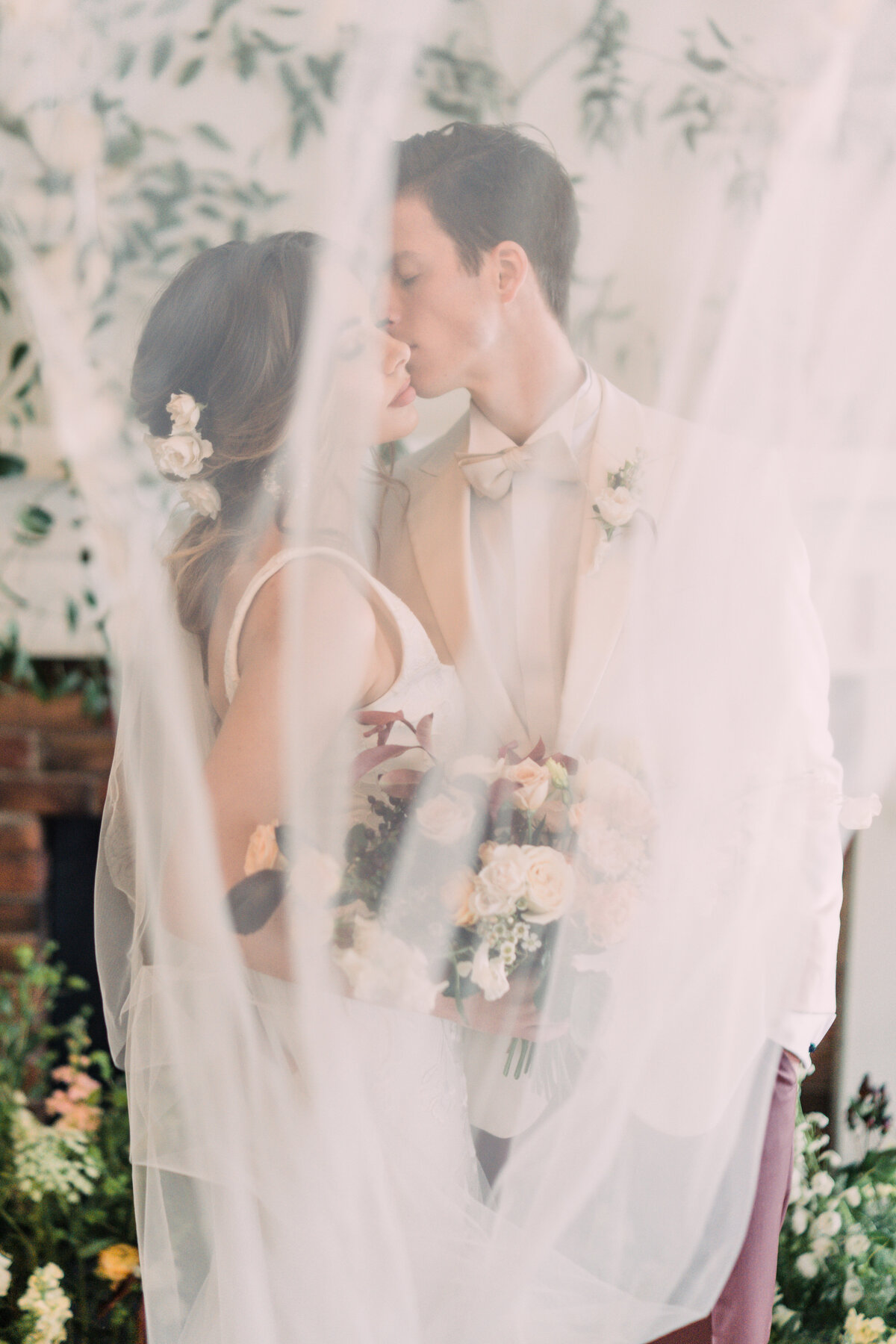 Veil covering bride with bouquet & flowers in her hair kissing groom in white tux with vines in background