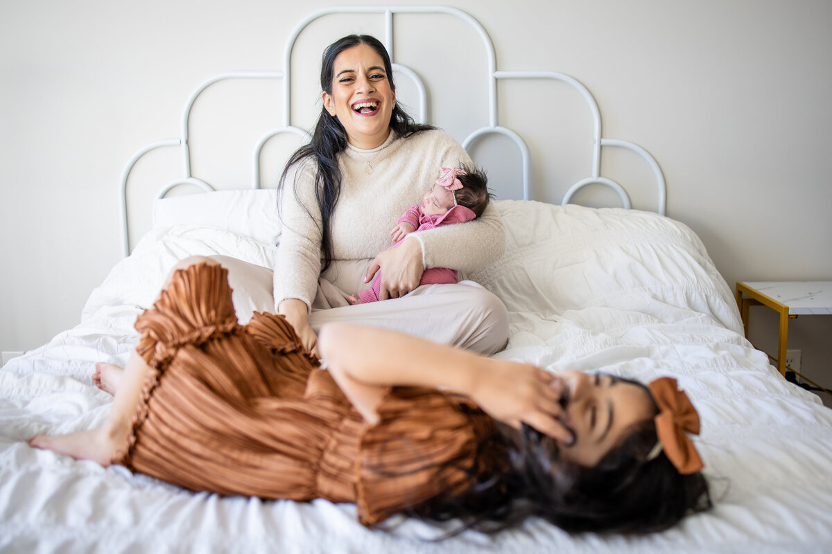 Mom sitting on bed in city apartment holding newborn daughter while her older daughter plays on the bed
