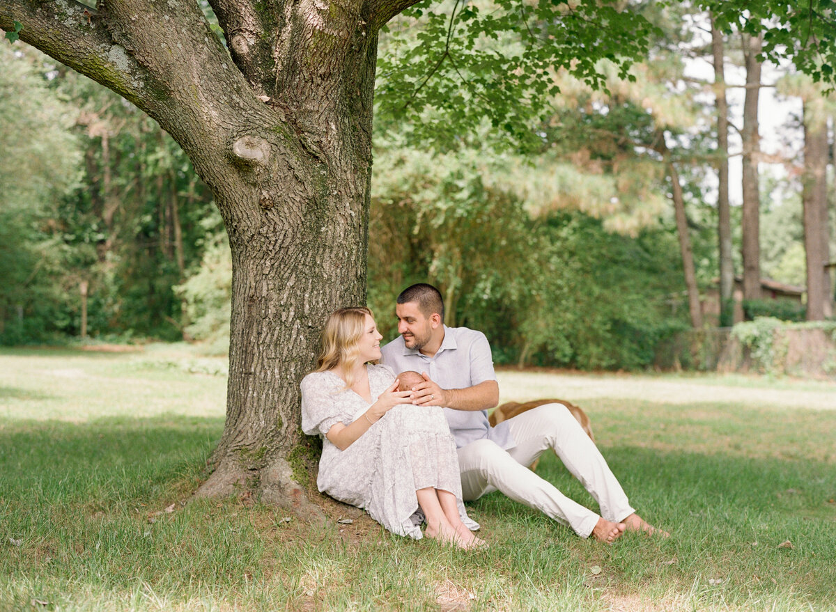 Family sits under a tree in their backyard and holds their newborn baby during a Raleigh NC newborn session. Photographed by Raleigh Newborn Photographer A.J. Dunlap Photography.