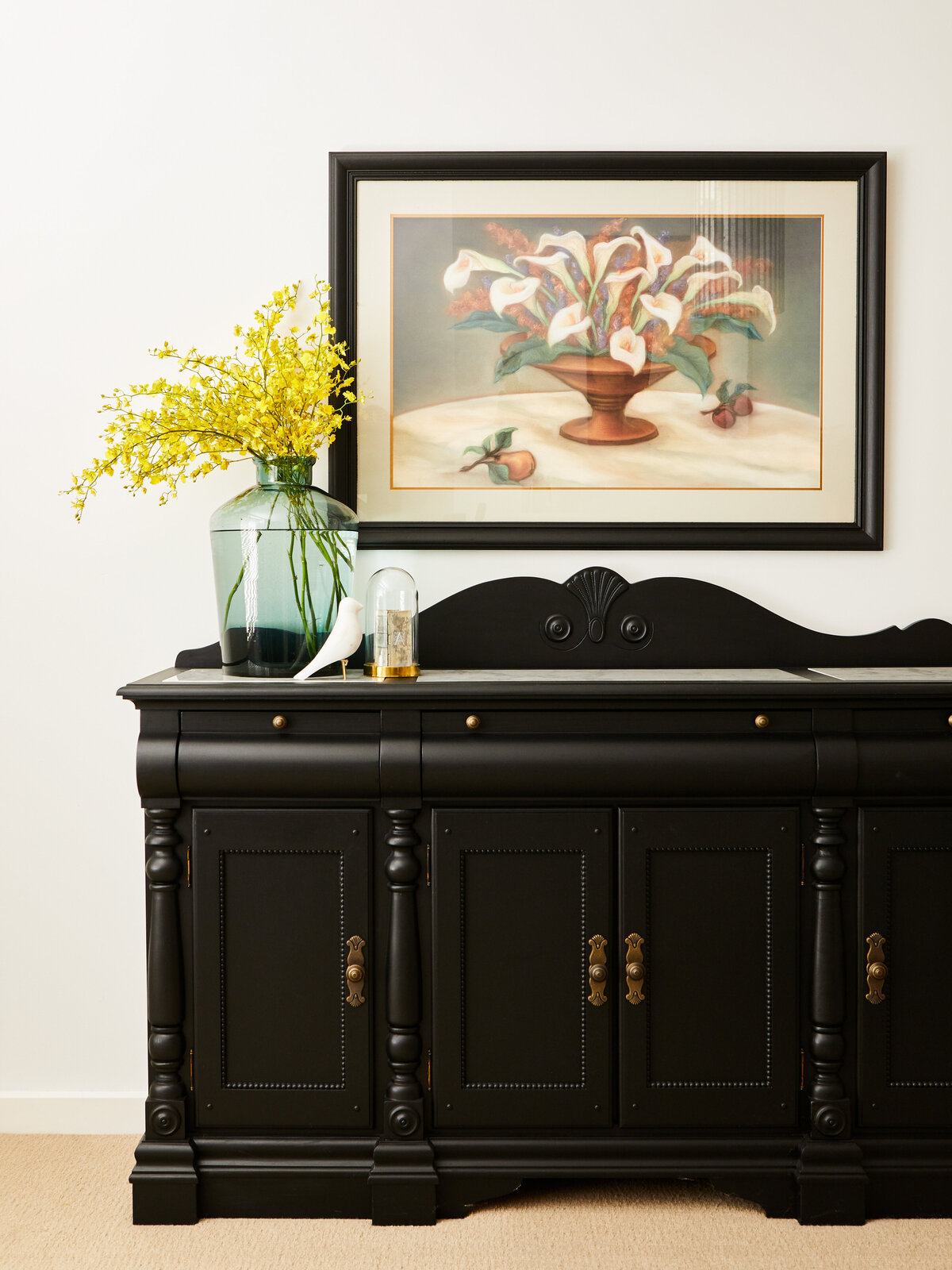 A black sideboard with a marble top and brass hardware in a home in Eynesbury. The surface is decorated with a turquoise glass vase of yellow flowers and a framed painting of white calla lilies.