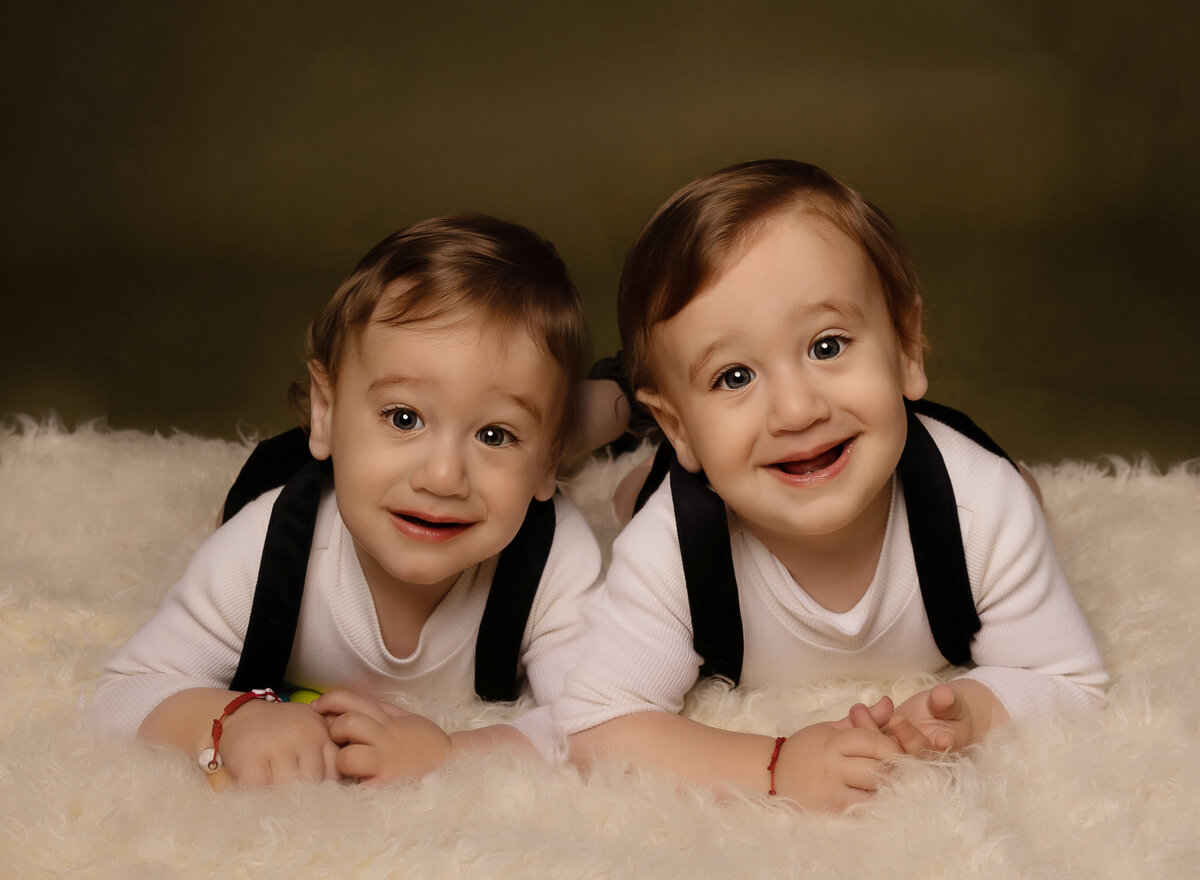 Twin baby boys pose for their Brooklyn NY first birthday photoshoot. the boys are laying on their belly with their heads touching and smiling at the camera. Captured by premier Brooklyn NY family photographer Chaya Bornstein Photography.