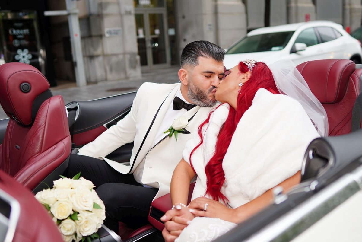 A couple enjoys a romantic kiss inside a convertible car, with the top down and the cityscape in the background. The image exudes a carefree and joyful vibe, highlighting the couple's affection and the fun of their special day.