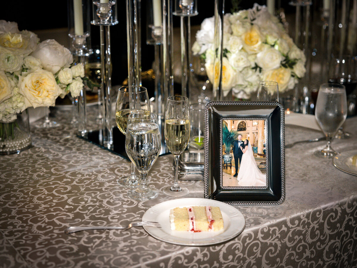 A wedding table setting with a slice of cake on a plate, two champagne glasses, floral centerpieces with white roses, and a framed photo of a couple in wedding attire. The tablecloth has a delicate pattern.