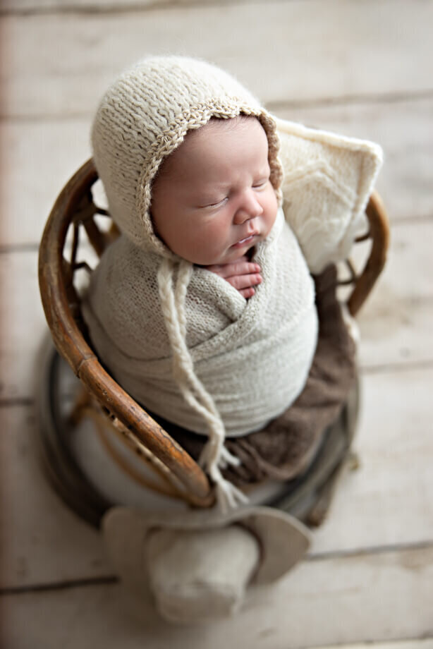newborn boy is swaddled in a cream wrap, wearing a matching knit beanie and posed in a wicker chair and decorated with a lasso.