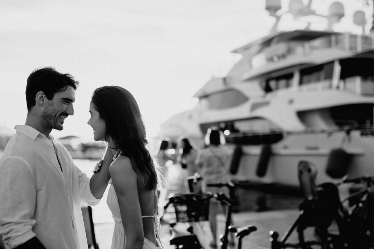 Black and white photo of the couple looking at each other and his arm on her face with a yacht in the background.