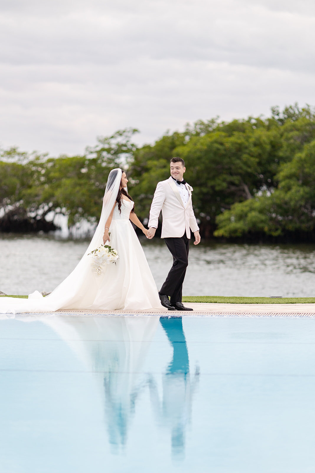 The bride and groom walking hand in hand by a serene pool, their reflection mirrored in the water, during their wedding at Ocean Reef in Key Largo, captured by Claudia Amalia Photography.