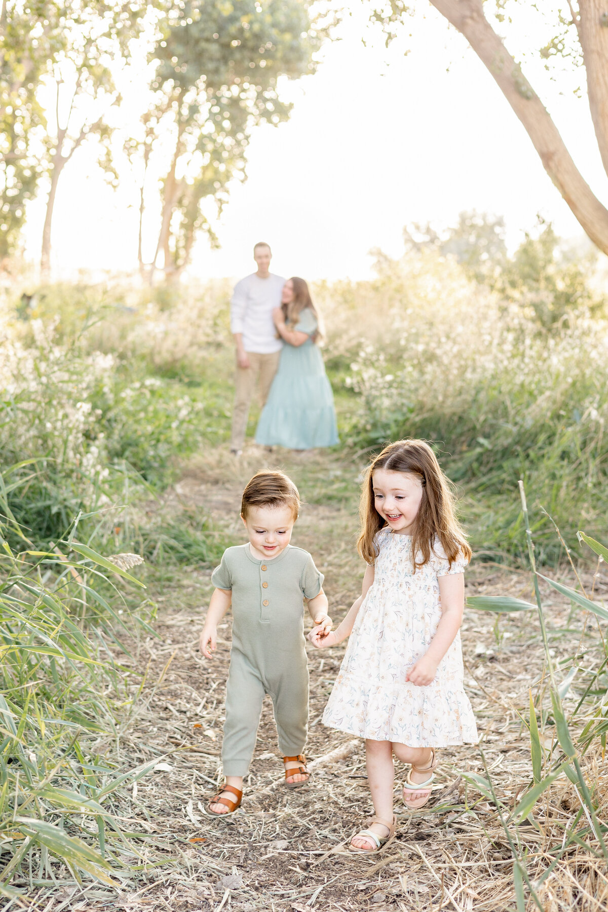 A young boy and girl holding hands and walking along a wooded path, with their parents blurred in the background.