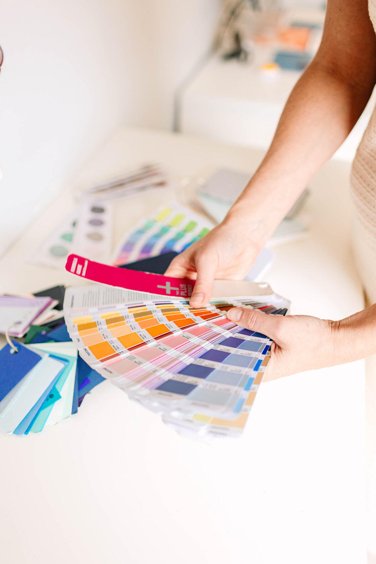 A person holds a fan deck of colorful Pantone swatches, selecting a shade of pink, with various color samples and design materials spread out on a white desk.