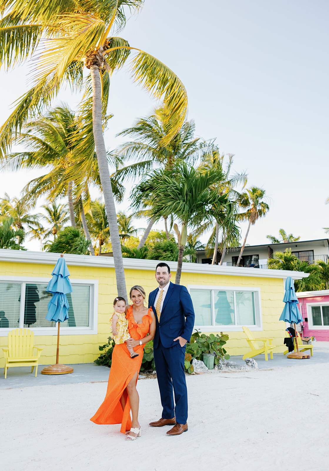 Wedding guests in bright outfits pose with their child against La Siesta Marina's vibrant yellow and pink beach cottages.