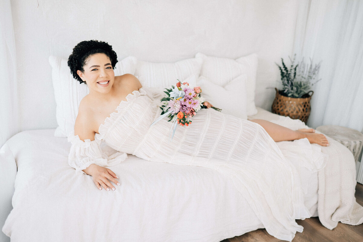 pregnant woman laying on a bed, holding flowers at her hip while looking at Denver photographer