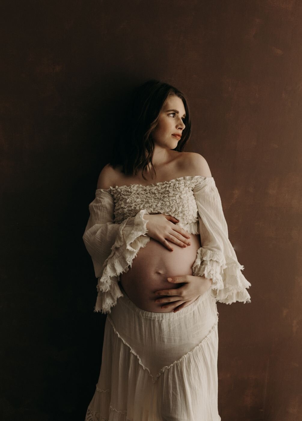 Elegant pregnant woman in a flowing white dress against a dark backdrop in Mullingar maternity photo