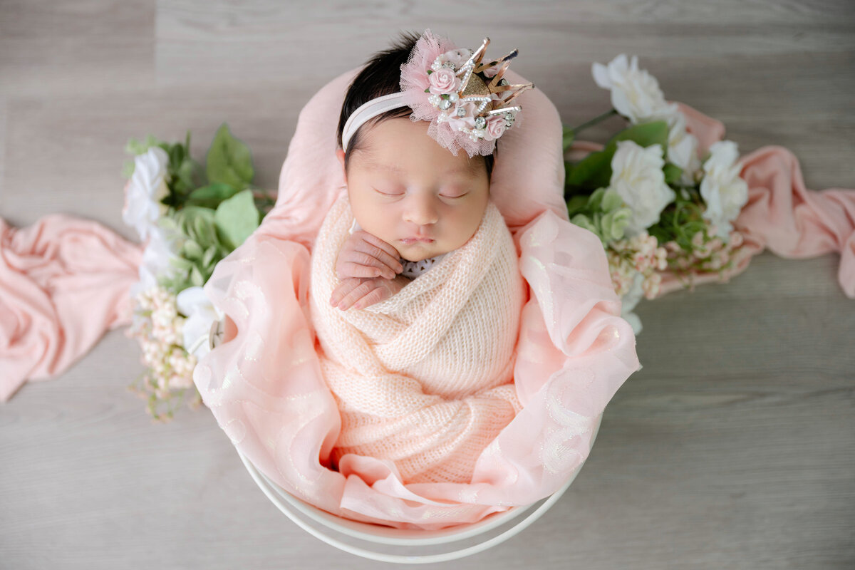 Newborn wrapped in peach fabric, with hands under chin inside a bucket, with soft floral accents on each side.