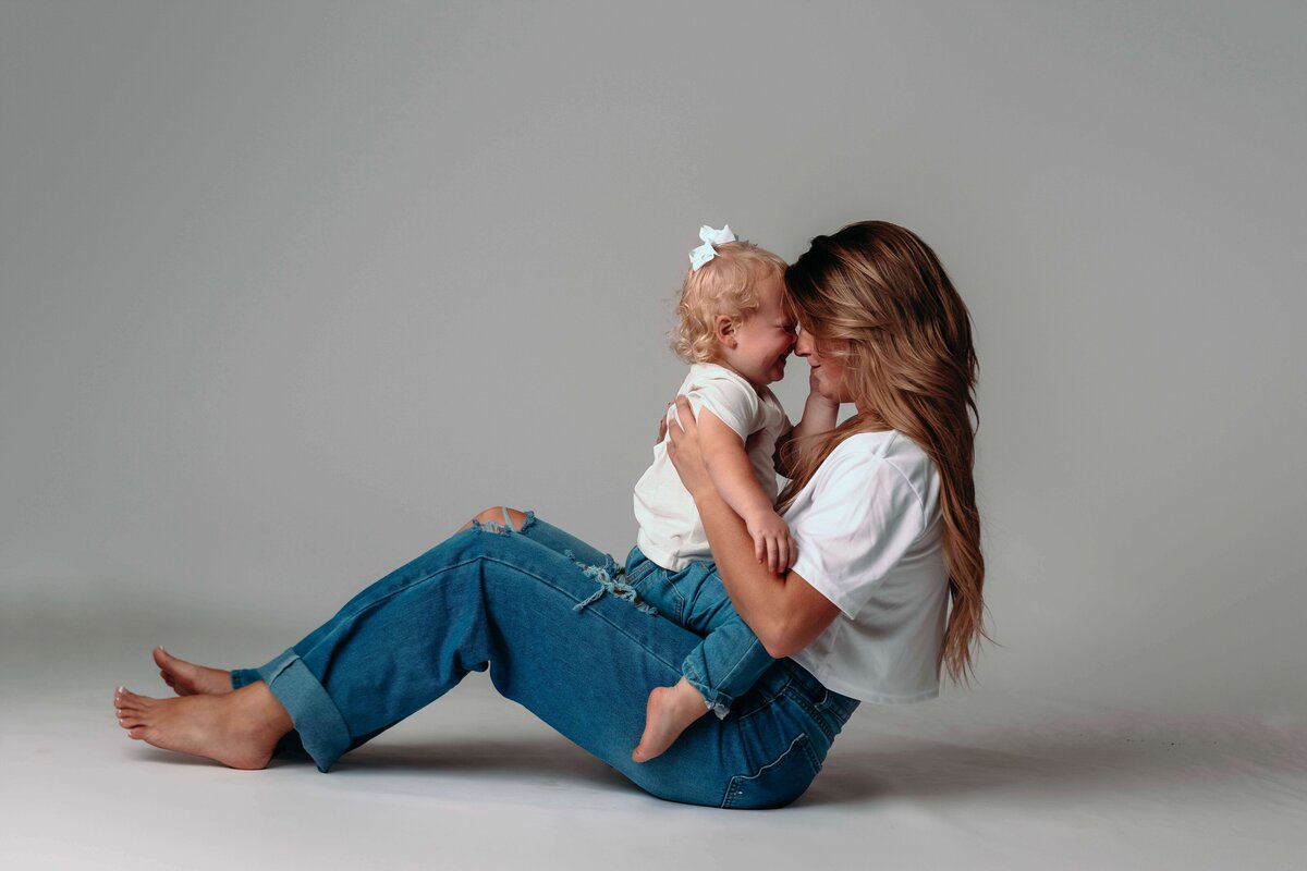 A young mom sits in the studio floor of Mobile, AL Family and Motherhood photographer, playing with her toddler aged daughter who is sitting in her lap. They are resting their foreheads together to show their closeness. They are sporting matching jeans and white shirts