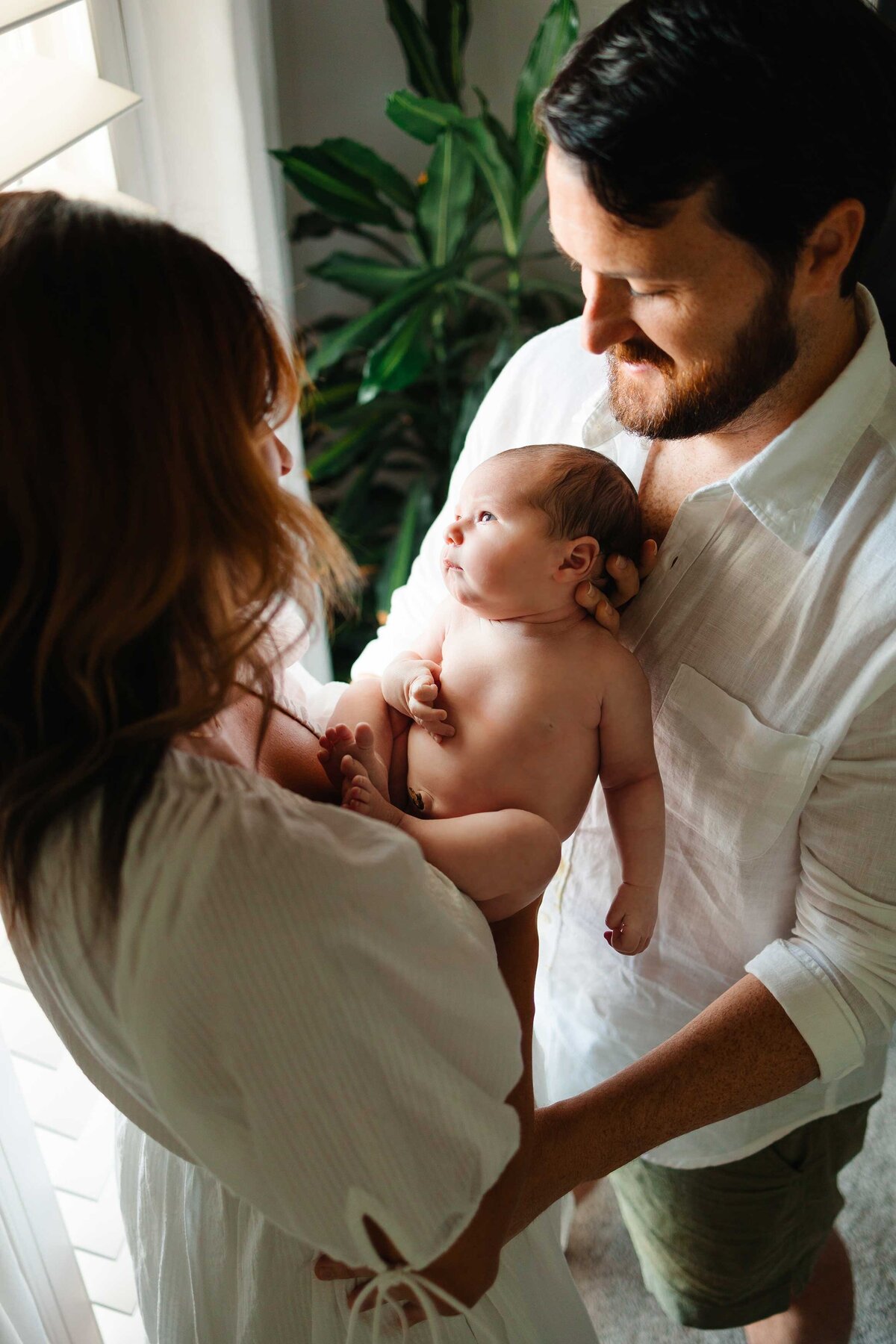 The joyful parents lovingly hold their newborn baby in front of a large window, adorned with a plant. They are dressed in stylish boho outfits, capturing a beautiful family moment.