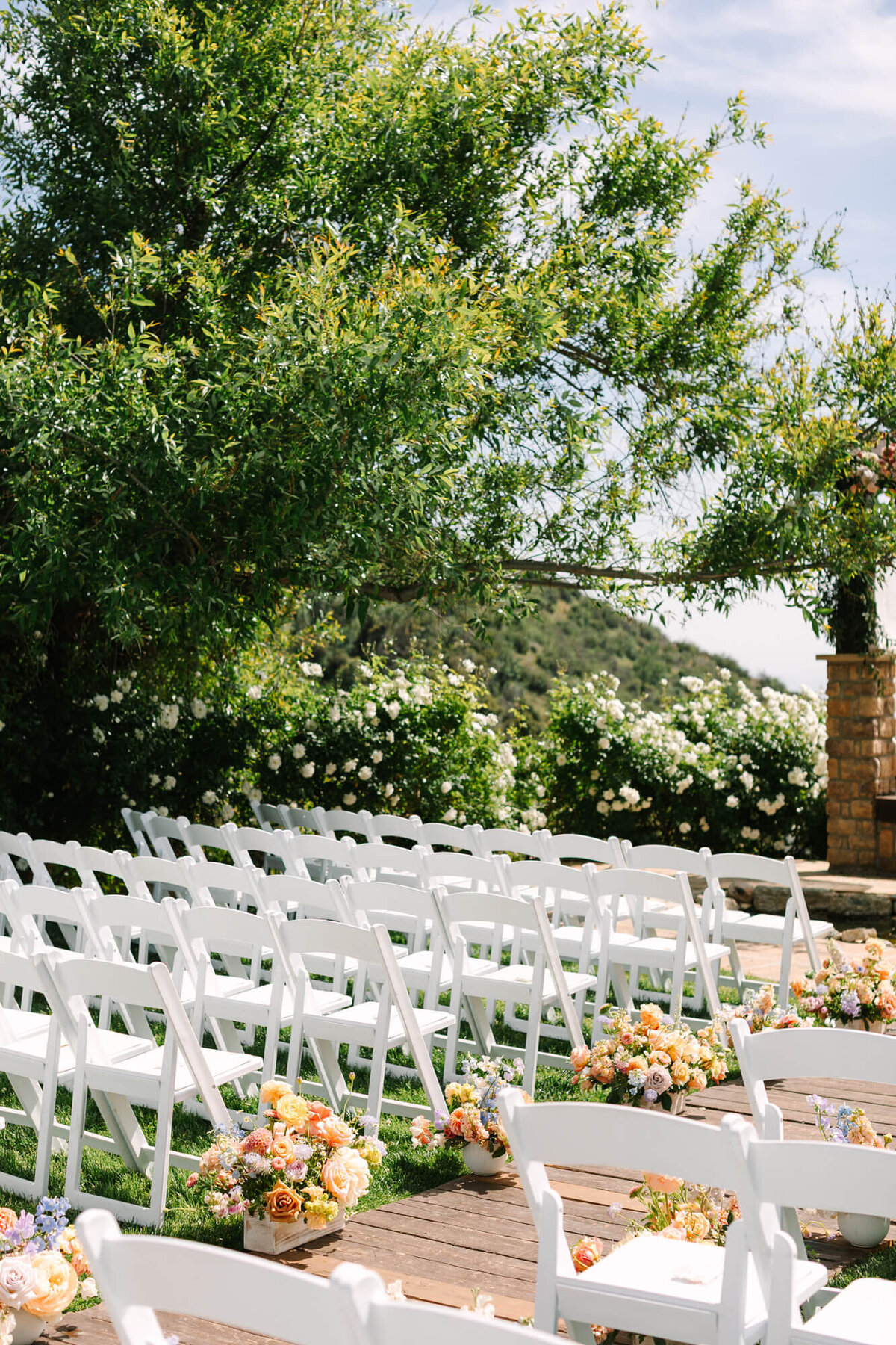 Wedding ceremony setup with rows of white chairs facing an altar. Colorful flower arrangements line the aisle. Lush greenery and a clear sky create a serene, festive atmosphere.