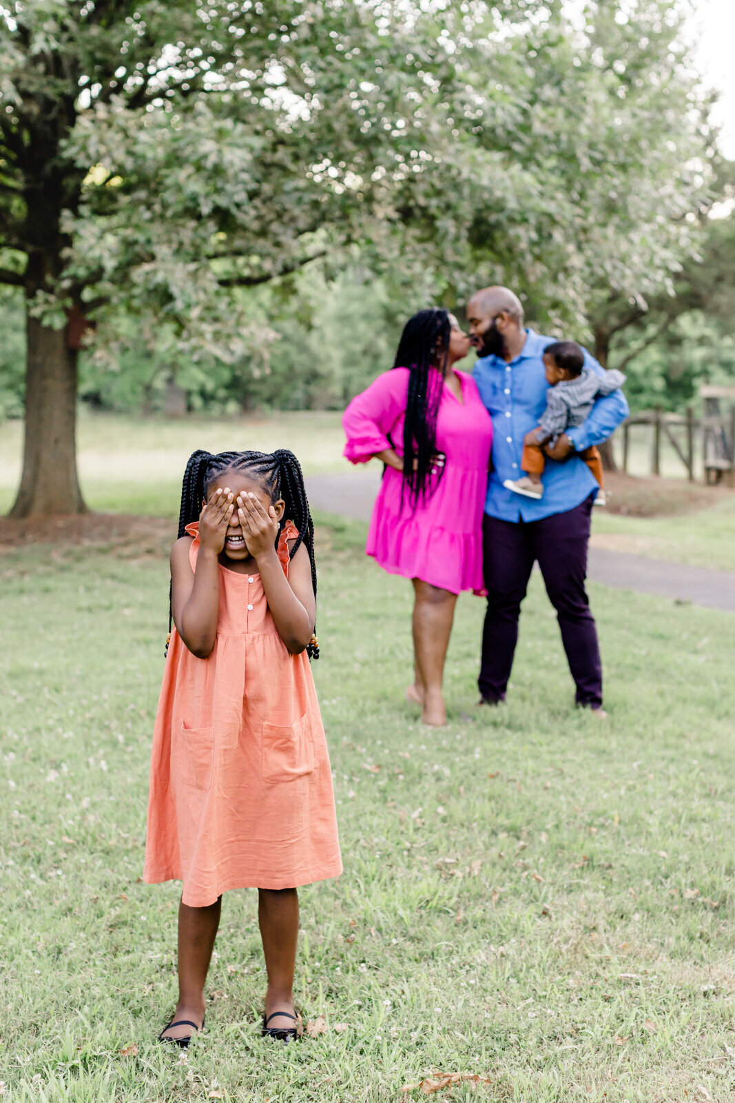 Young daughter covers her eyes while standing in front of parents who are kidding while holding their young son at McDaniel Farm Park