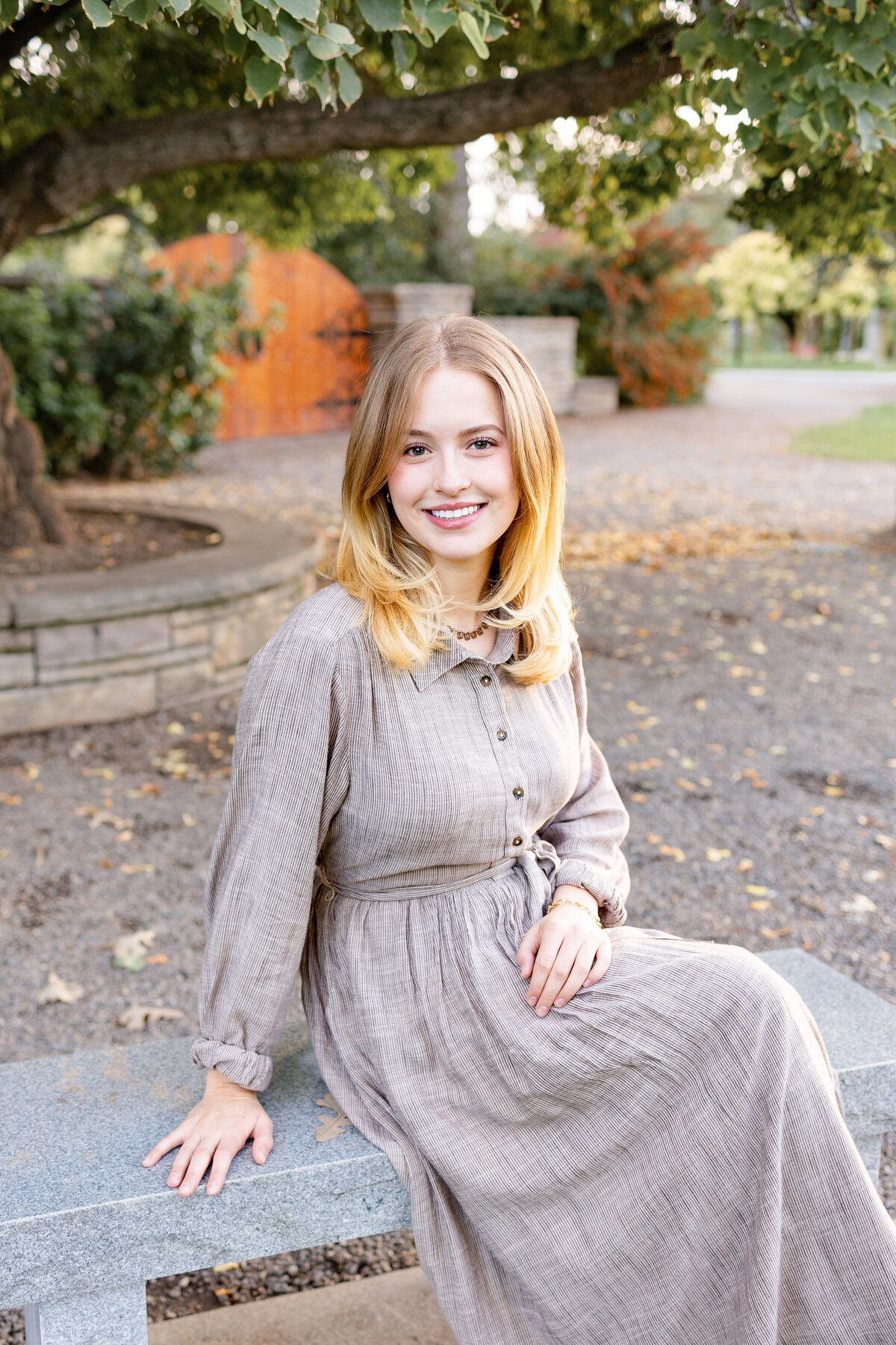 A young woman sits gracefully on a stone bench under a tree, smiling softly as she enjoys the peaceful surroundings.