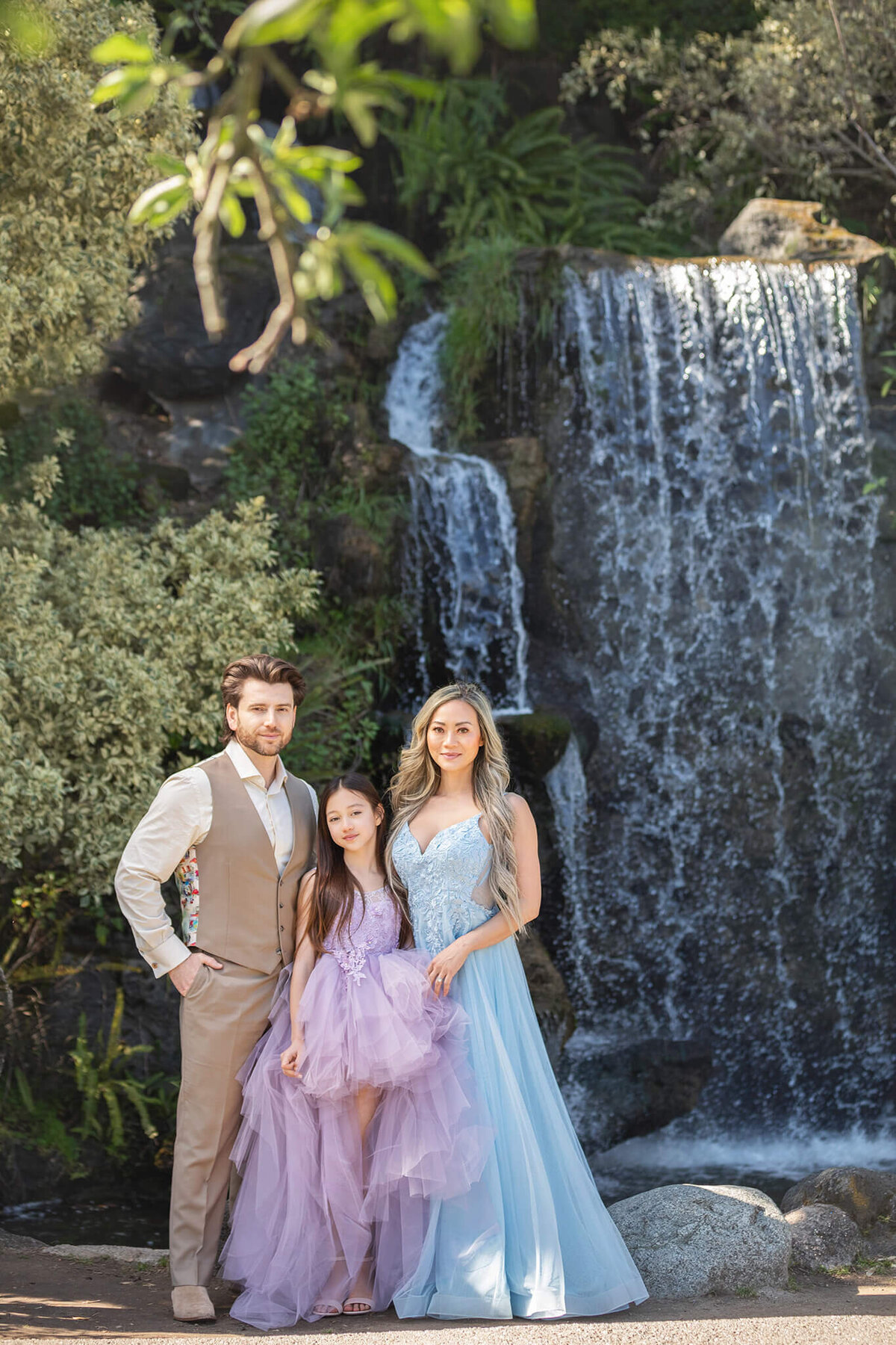 Mom and daughter in beautiful long gowns and dad in a beige suit photographed in front of a waterfall at Los Angeles Arboretum - Los Angeles Family Photography