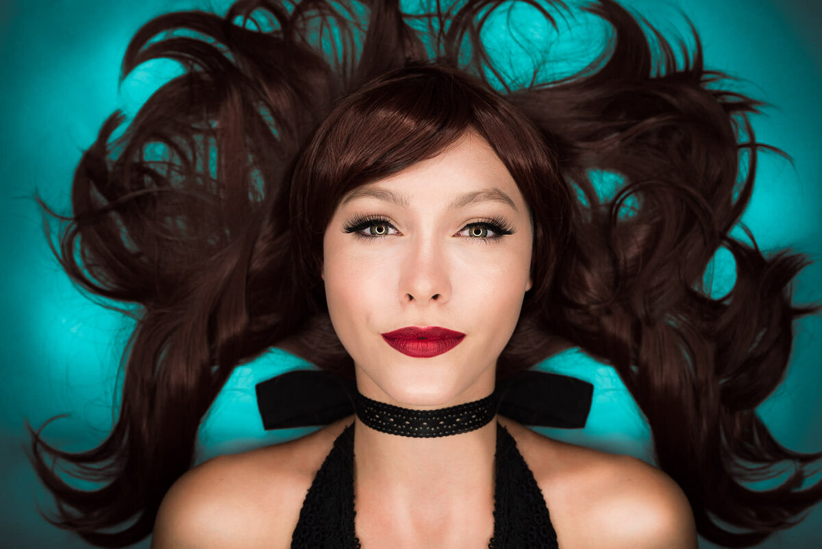 Creative overhead portrait of a woman with dark red hair fanned out in all directions against a bold bluish-green backdrop, photographed in Austin, TX.