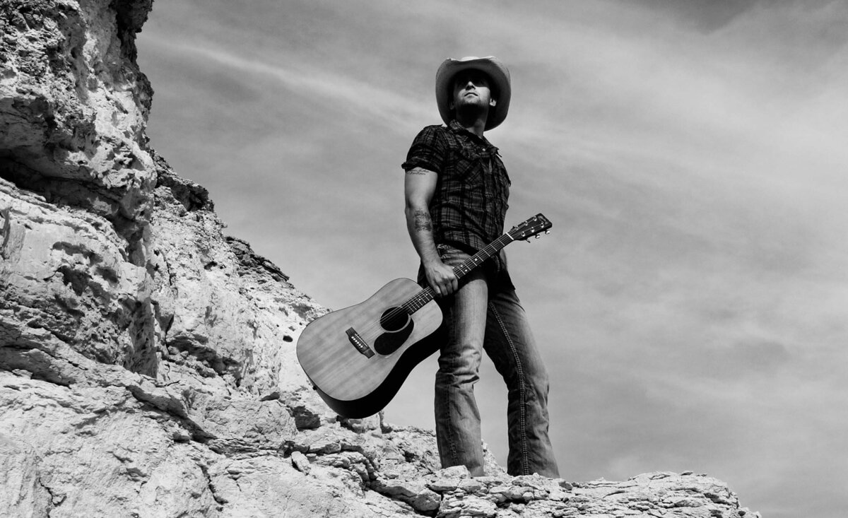 Country musician portrait Dean Brody black and white holding guitar wearing jeans straw cowboy hat standing against rock cliff