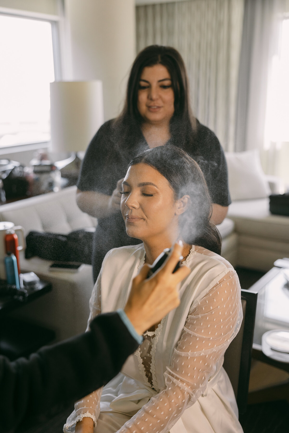 A makeup team sprays different sprays onto a bride sitting in a chair as she finishes her getting ready services.