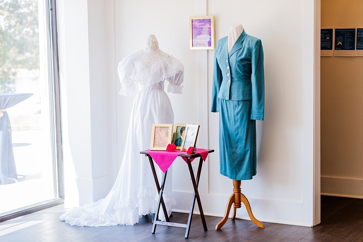 The bride and groom's grandmother's wedding dresses on display at Sugarfoot wedding. One dress has a long white train with elbow length puffed sleeves and a high neckline. The other dress is a long sleeved tea length teal suit. Photos of the brides are framed and sitting on a table with a hot pink accent.