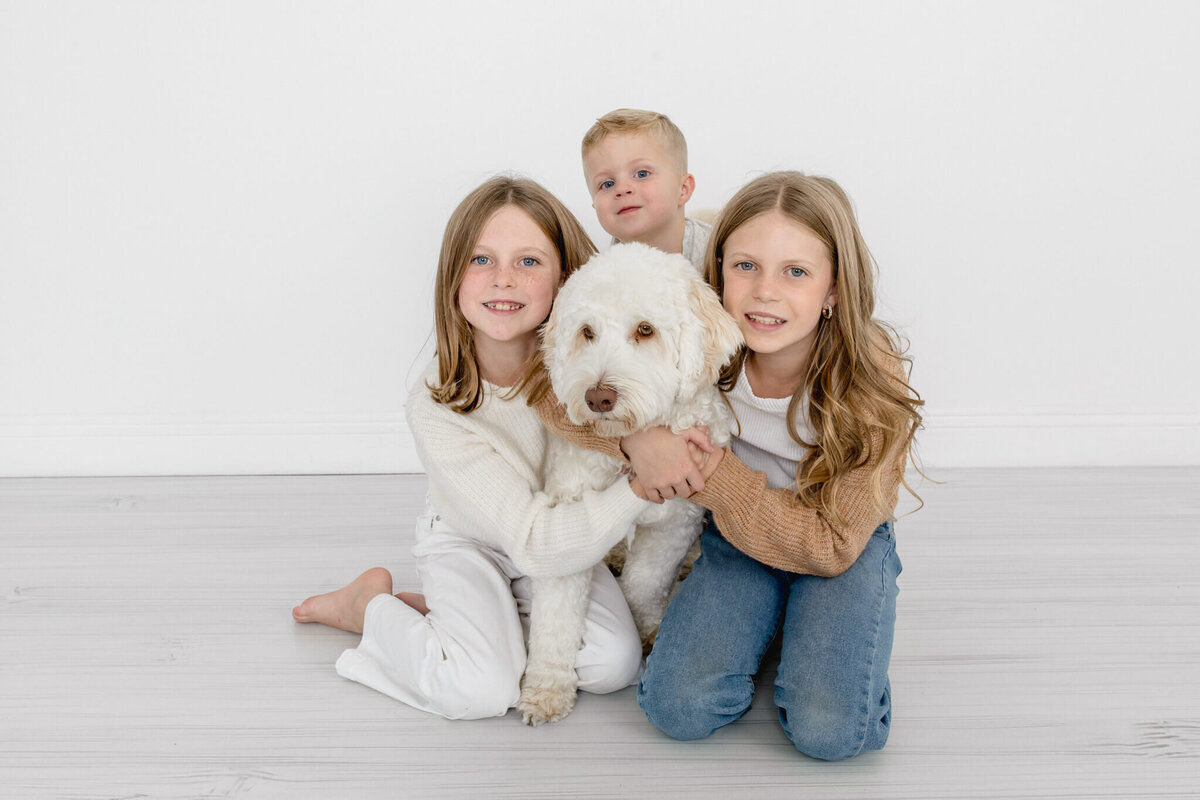 Three young kids with blonde hair sitting on a grey floor hugging their big, white fluffy dog and looking at the camera against a white wall.