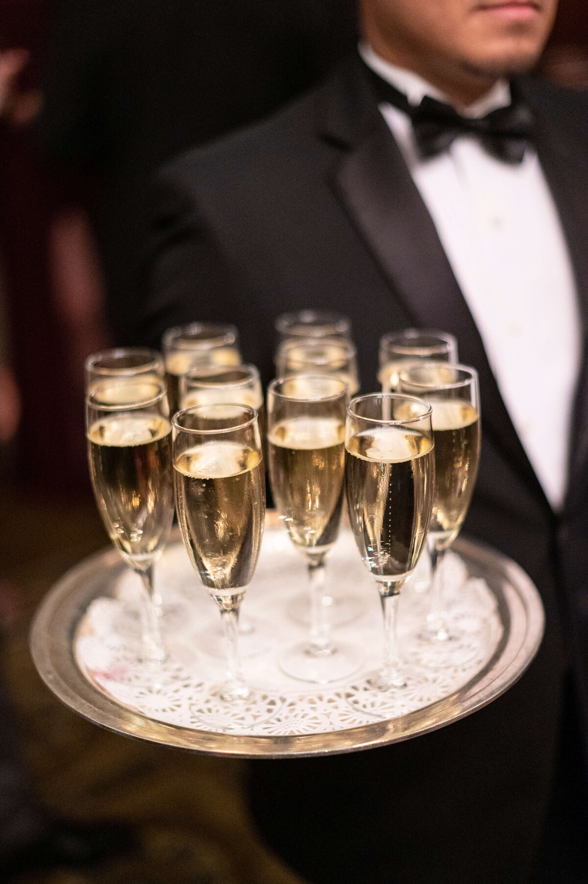 A person in a black tuxedo holds a silver tray with several filled champagne flutes, ready to be served at an elegant event.