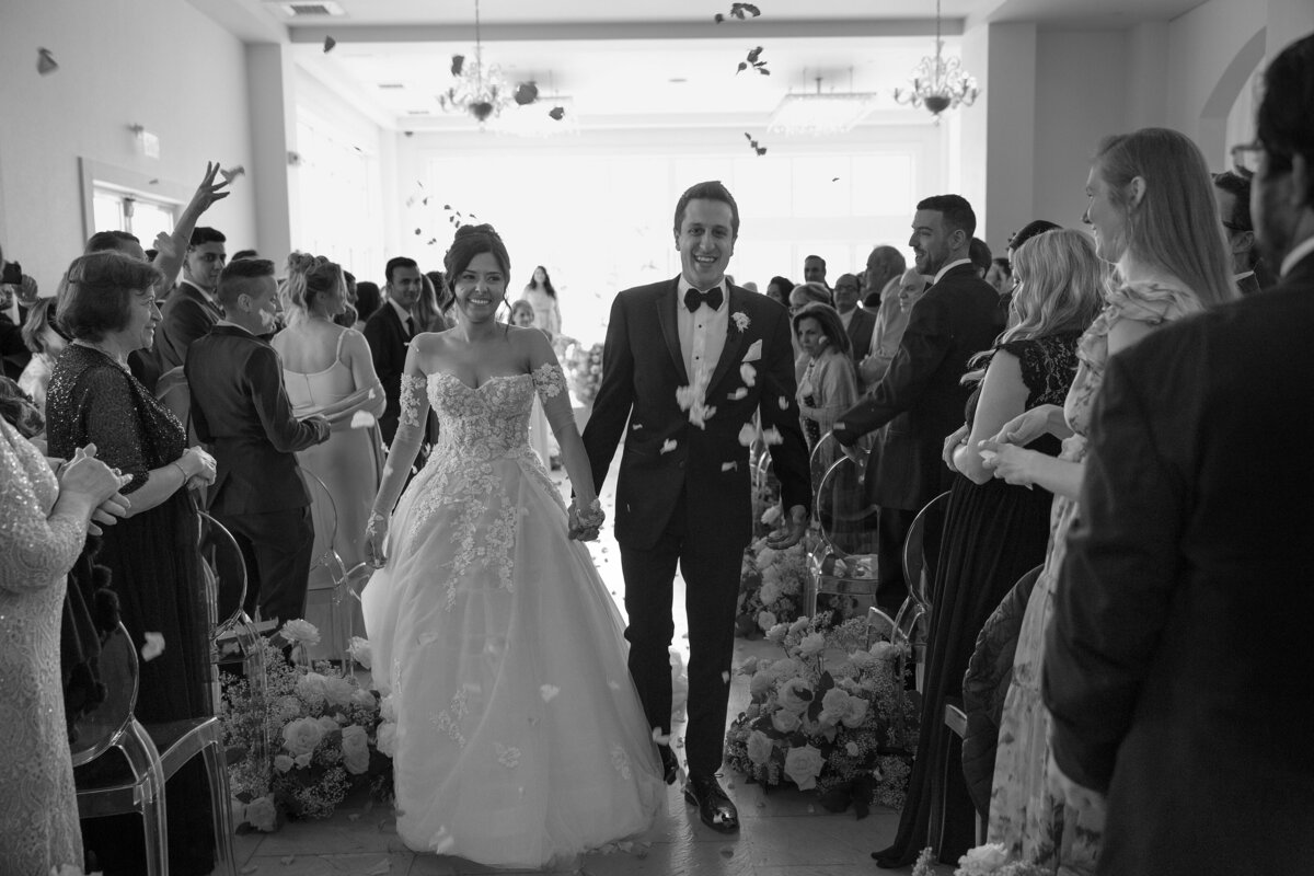 Bride and Groom smile as they walk down the aisle while guests throw rose petals.