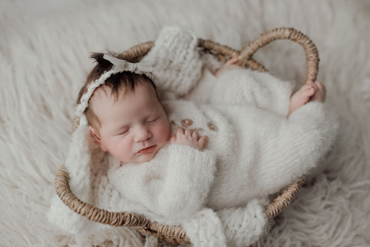 Studio newborn photography - baby girl sleeping in a basket wearing a cream knit onesie and lace headband. Her hand is resting gently on her chest