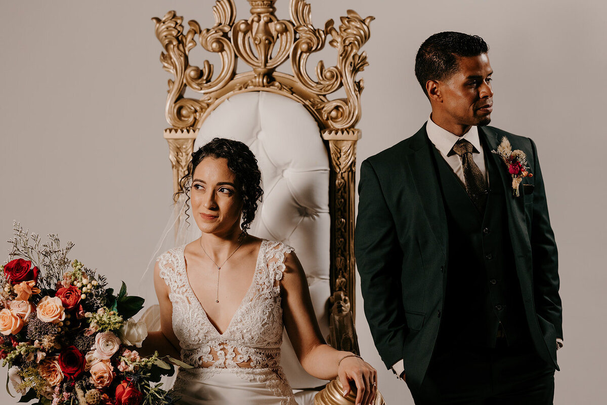The bride and groom pose next to a grand white and gold chair; she holds a vibrant bouquet, and he stands beside her in a green suit with a serious expression.