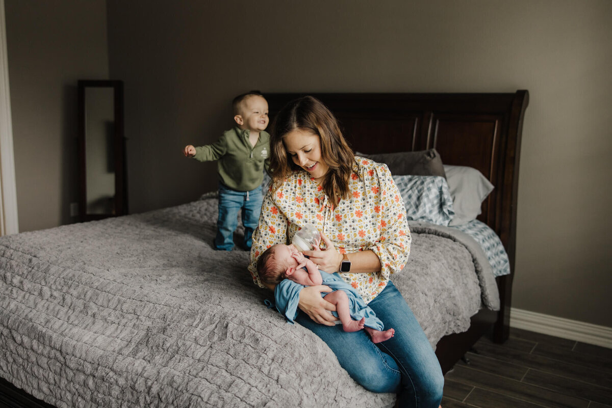 lifestyle newborn photo of mother feeding infant son in Longview, TX home while toddler jumps on bed