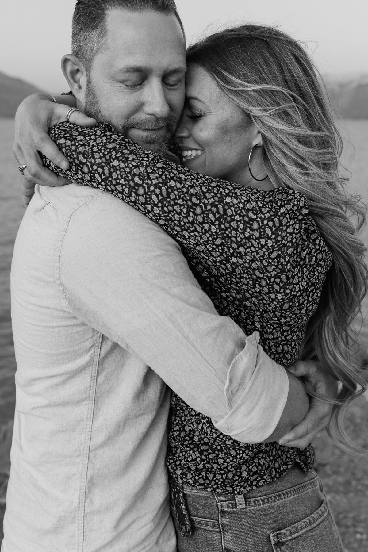 A black and white photograph of a couple embracing near Utah Lake at the Saratoga Springs Marina. The woman smiles with joy as she wraps her arms around her fiancé’s shoulders, their love captured in a timeless and intimate moment.