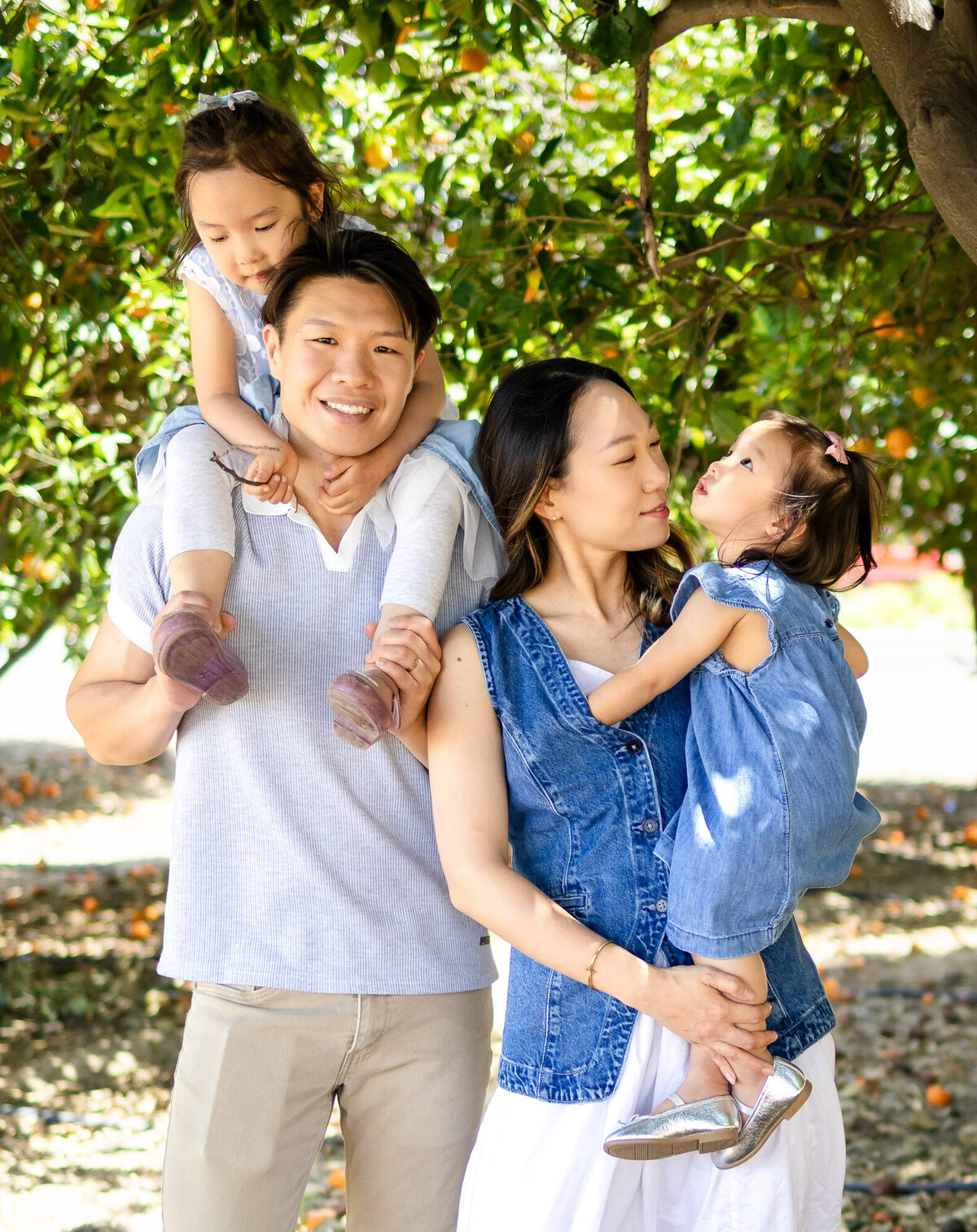 Bay Area Family Portfolio - Ellobelle Photography highlights parents smiling while carrying their daughters, framed by orange trees and natural light.