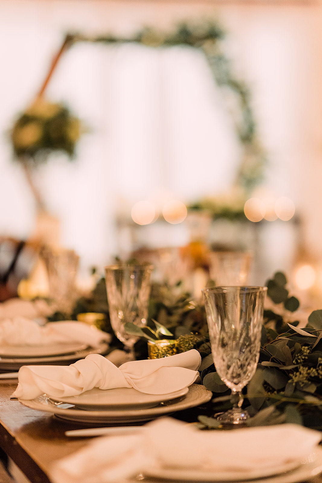 A close-up of an elegantly decorated wedding reception table with folded napkins, crystal glasses, and greenery centerpieces.