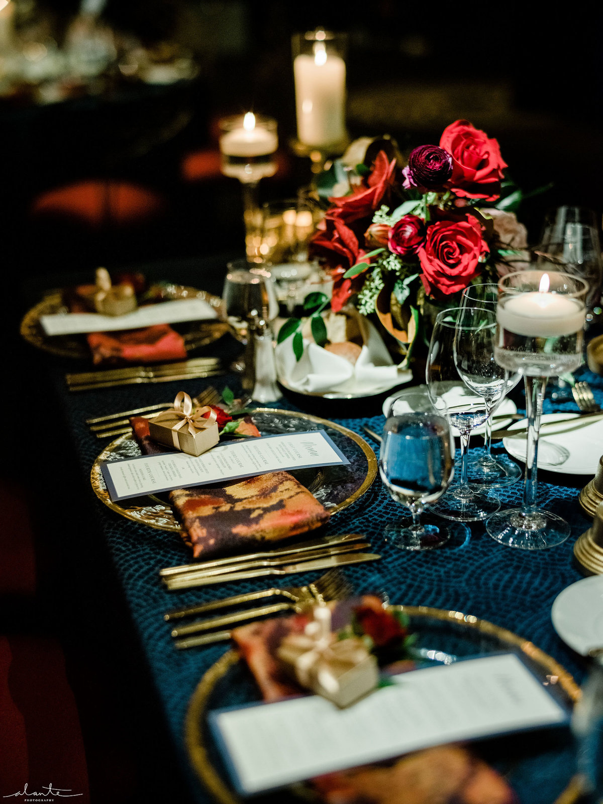 table scape at wedding reception with red roses, gold candle holders, and red and gold napkins on blue linen