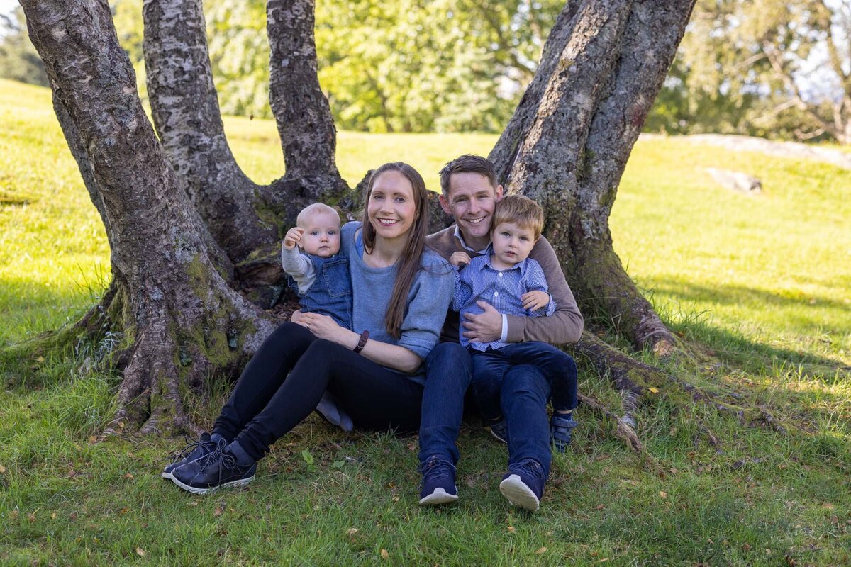 Familiefoto i park. Familien sitter ved fotenden til noen trær, mor har minstemann på fanget og far har storebror på fanget. Fotografert i Langhaugparken av Fotograf Linn Landmark.