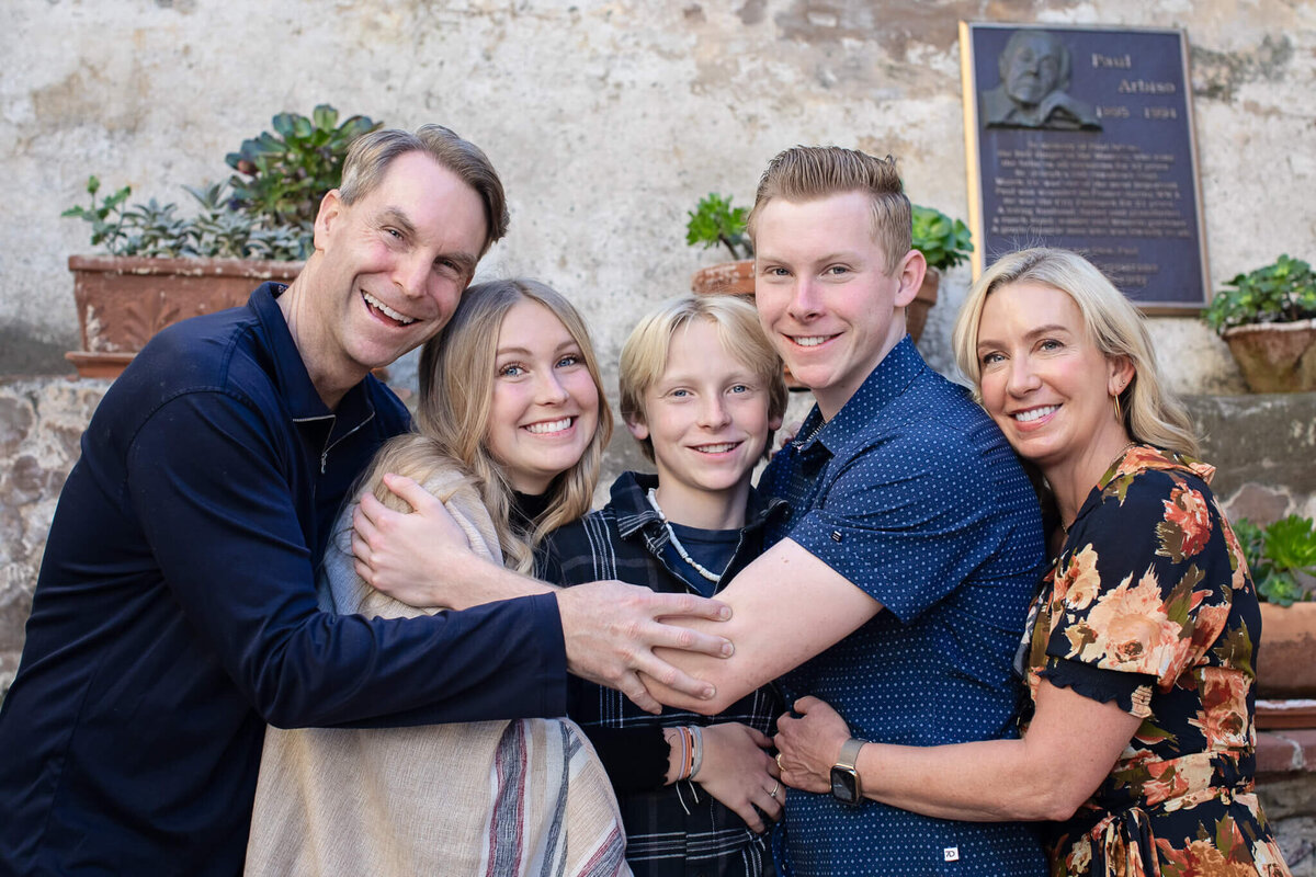 A blonde family of five hugging each other with their arms around one another, smiling at the camera.