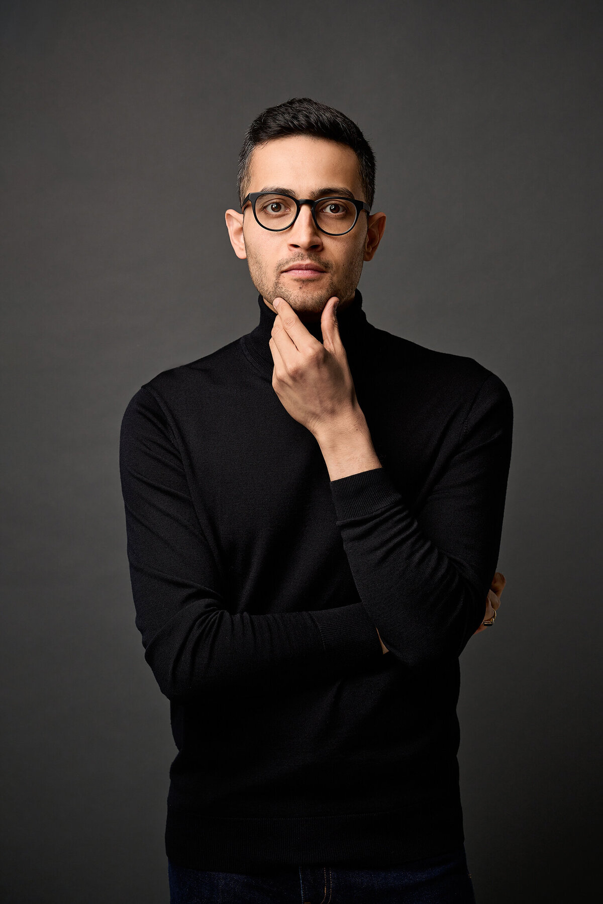 Man in black turtleneck and glasses with his hand on his chin.  He is looking intensively at the camera. The tones are moody against a dark grey background.