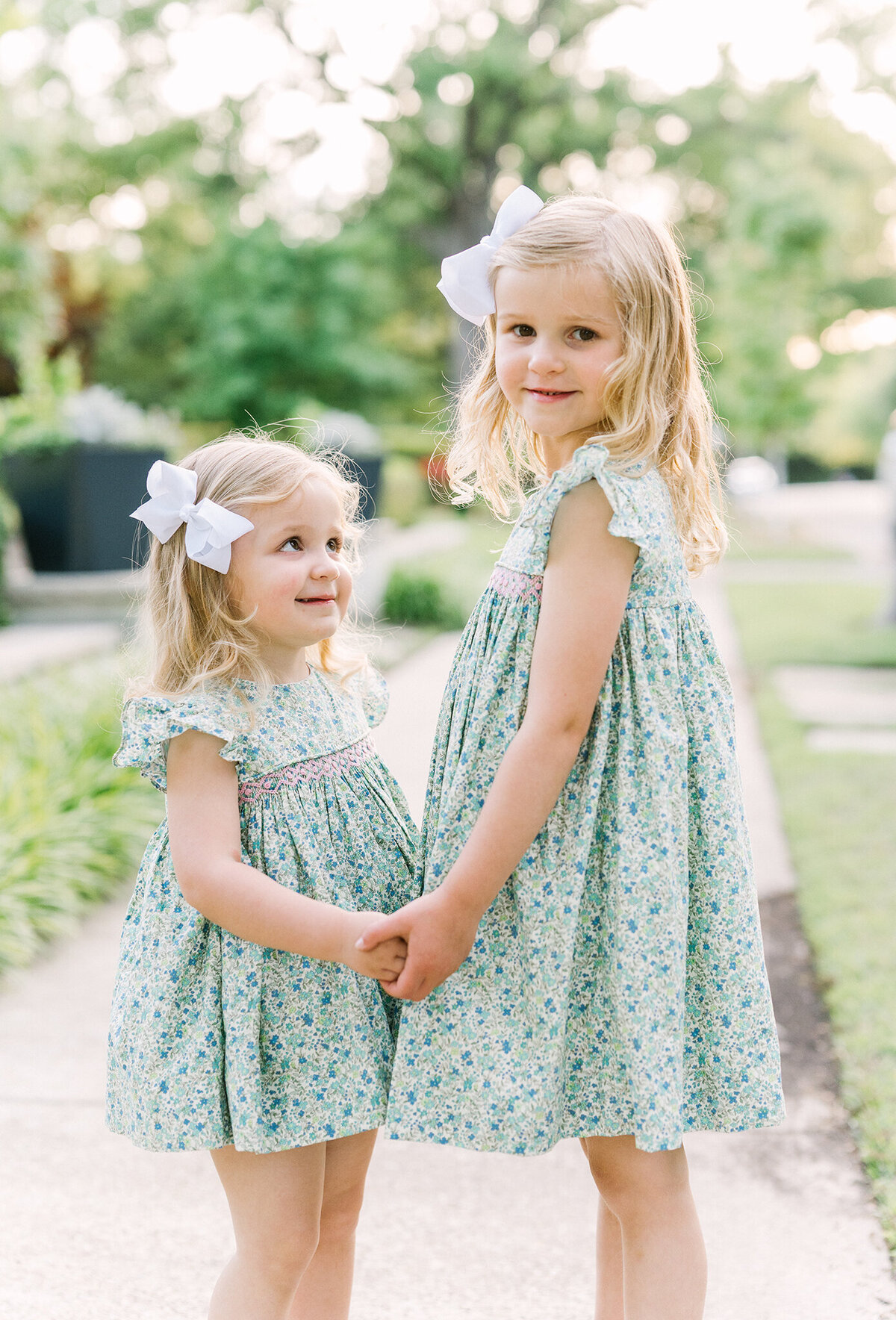 Sisters facing each other holding hands while wearing green floral smocked dresses on a sidewalk in Dallas.
