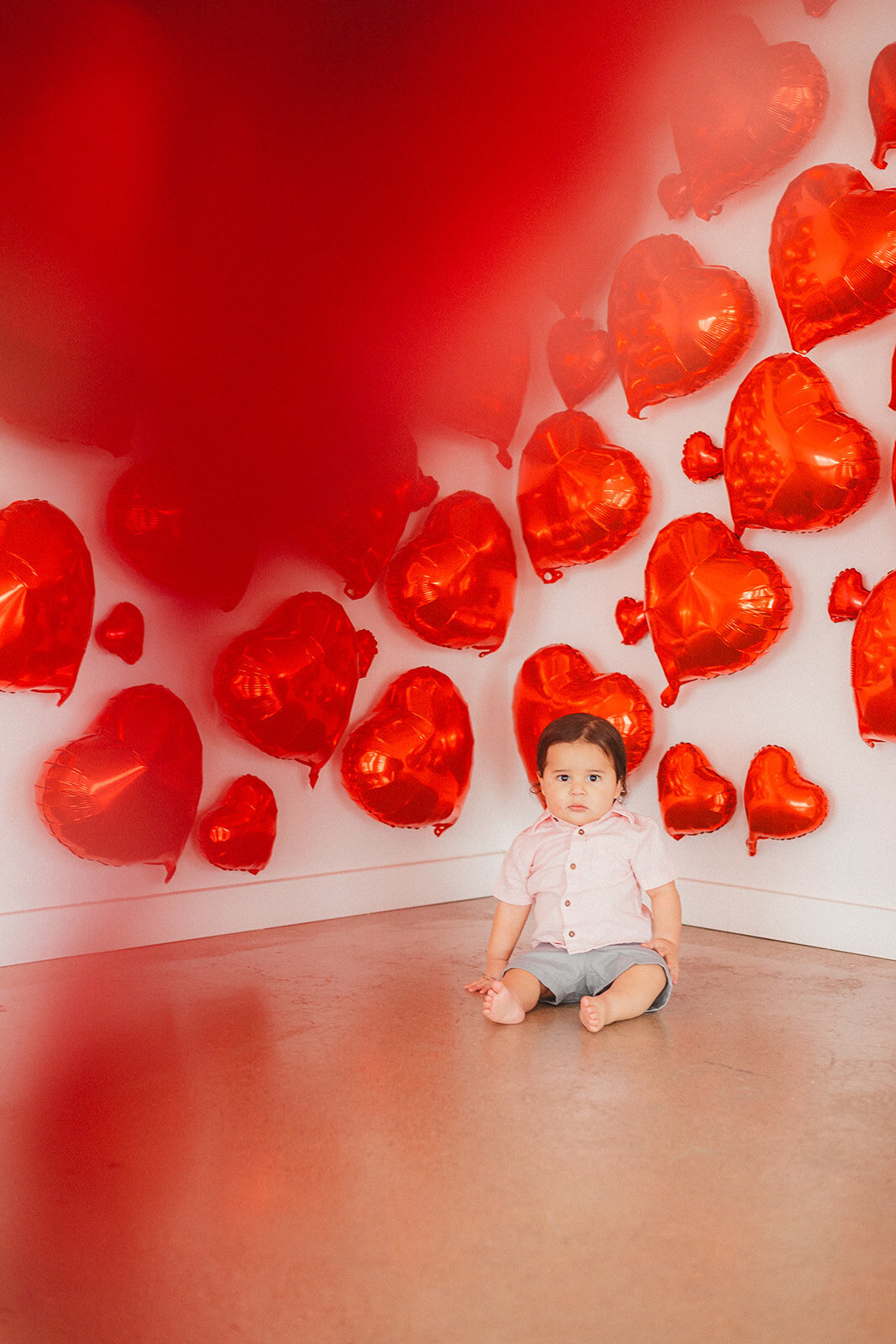 A baby boy stands in front of red heart balloons.