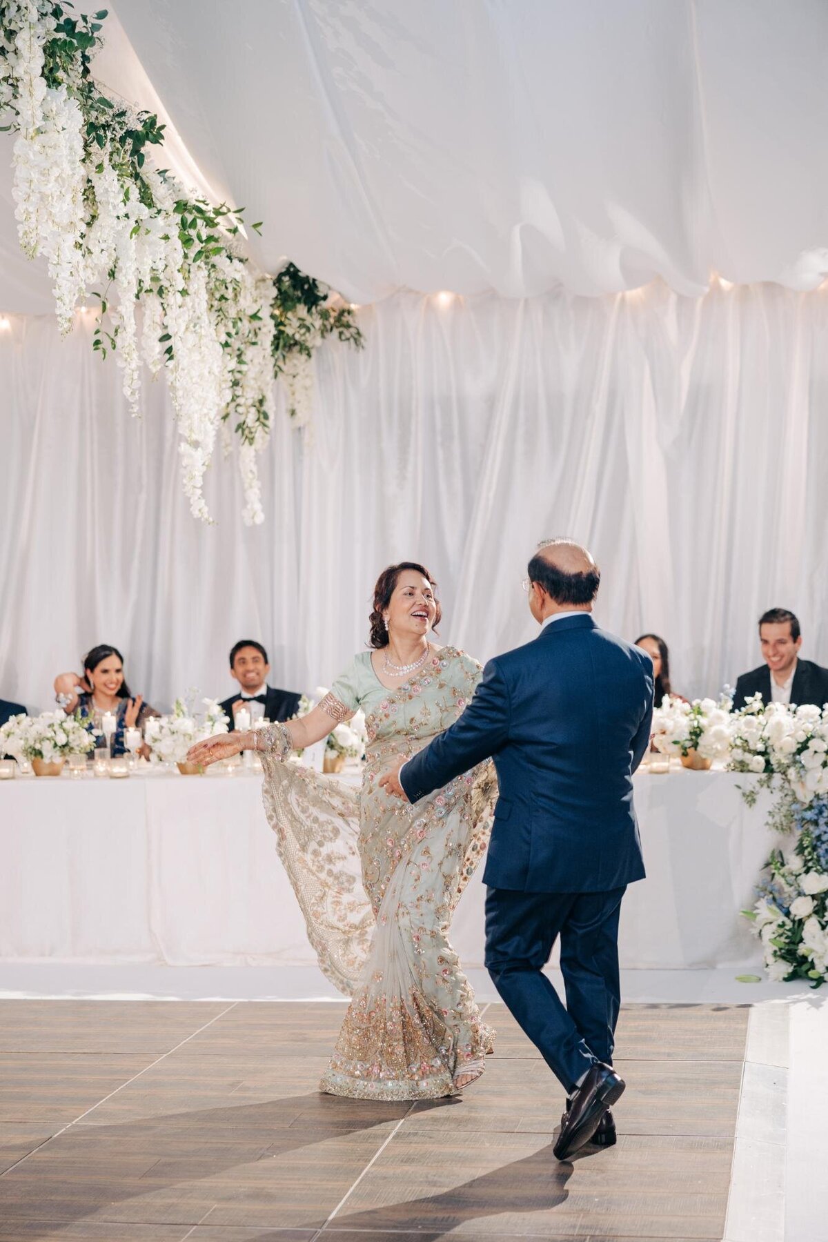 A bride in a beaded dress laughing joyfully while dancing with a man at a wedding reception, with guests watching in the background.