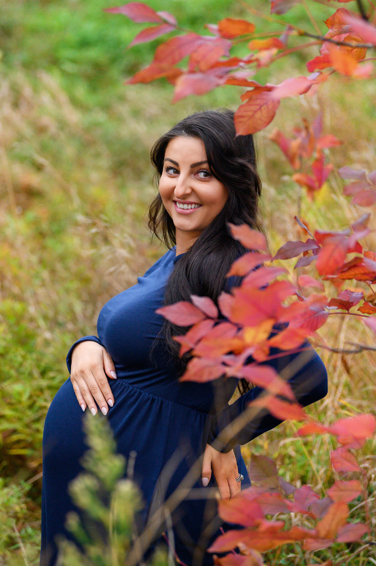 pregnant woman wearing a blue maternity dress holding her belly with a red leaved plant to the right for her fall outdoor maternity photography session in Burlington, Ontario 