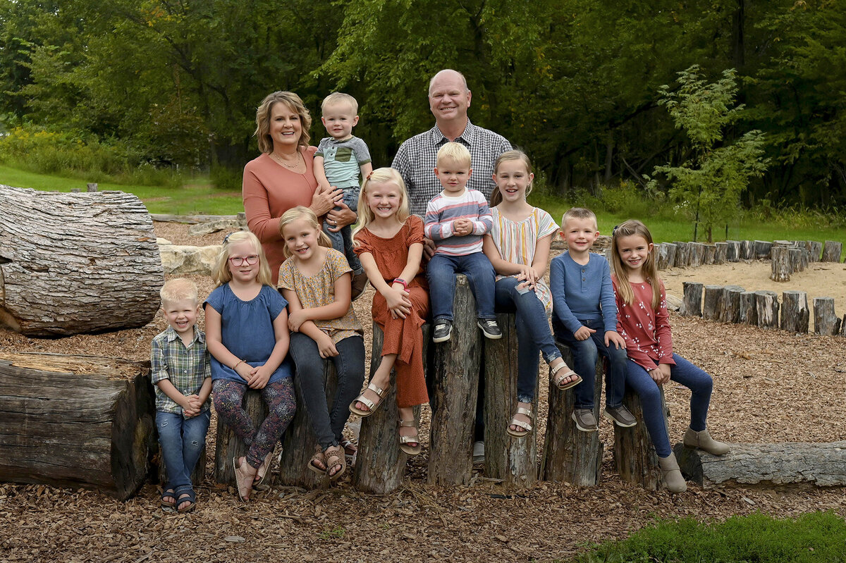 Family portrait outdoors with 2 adults and 9 children sitting on wooden logs, surrounded by trees and greenery