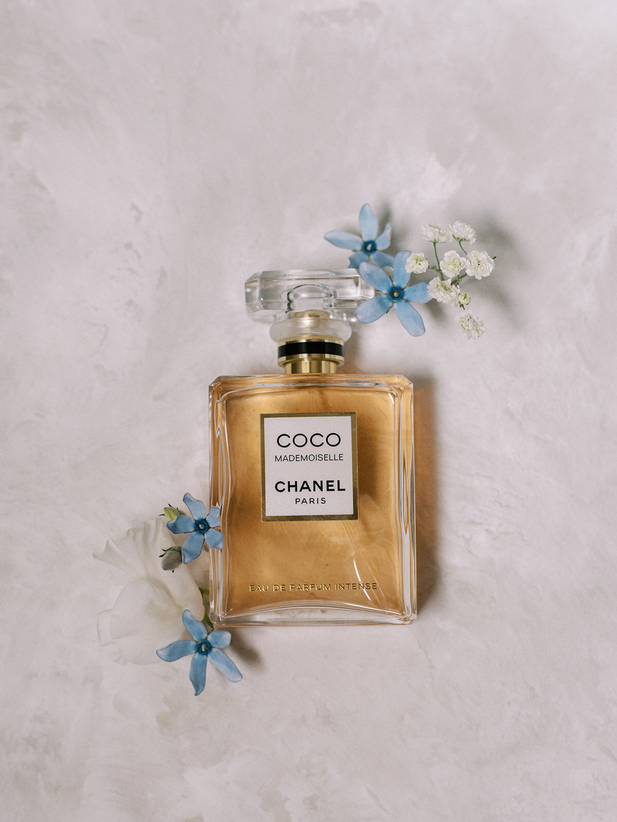 A Coco Mademoiselle Chanel perfume bottle placed on a light-colored textured surface, surrounded by small blue and white flowers, adds a touch of elegance to the scene. Perfect for capturing the essence of a classic Calgary wedding.